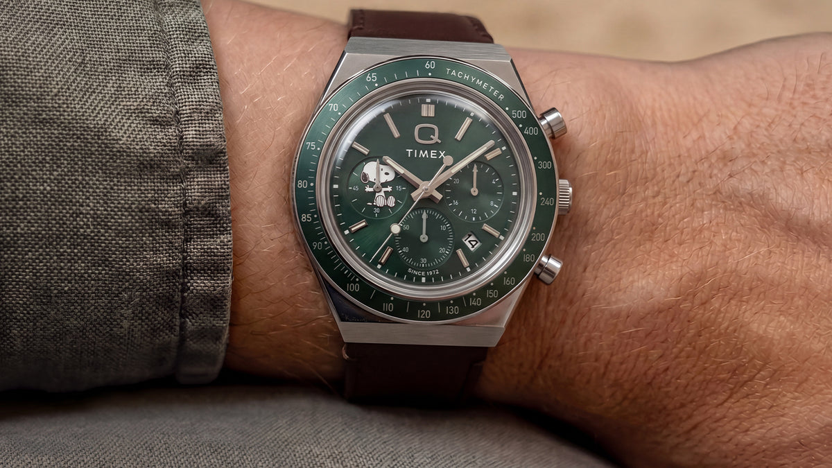 Primer plano del cronógrafo Peanuts® x Q Timex, con esfera verde y correa de piel marrón, puesto en la muñeca.