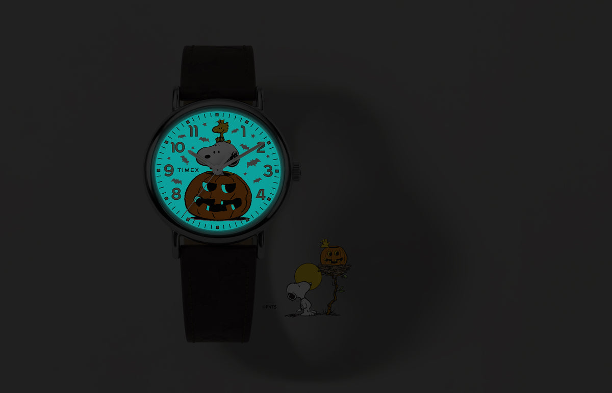 Peanuts® x Timex Weekender® Pumpkin watch shown in the dark with a glowing blue INDIGLO® dial revealing a bat pattern around Snoopy and Woodstock.