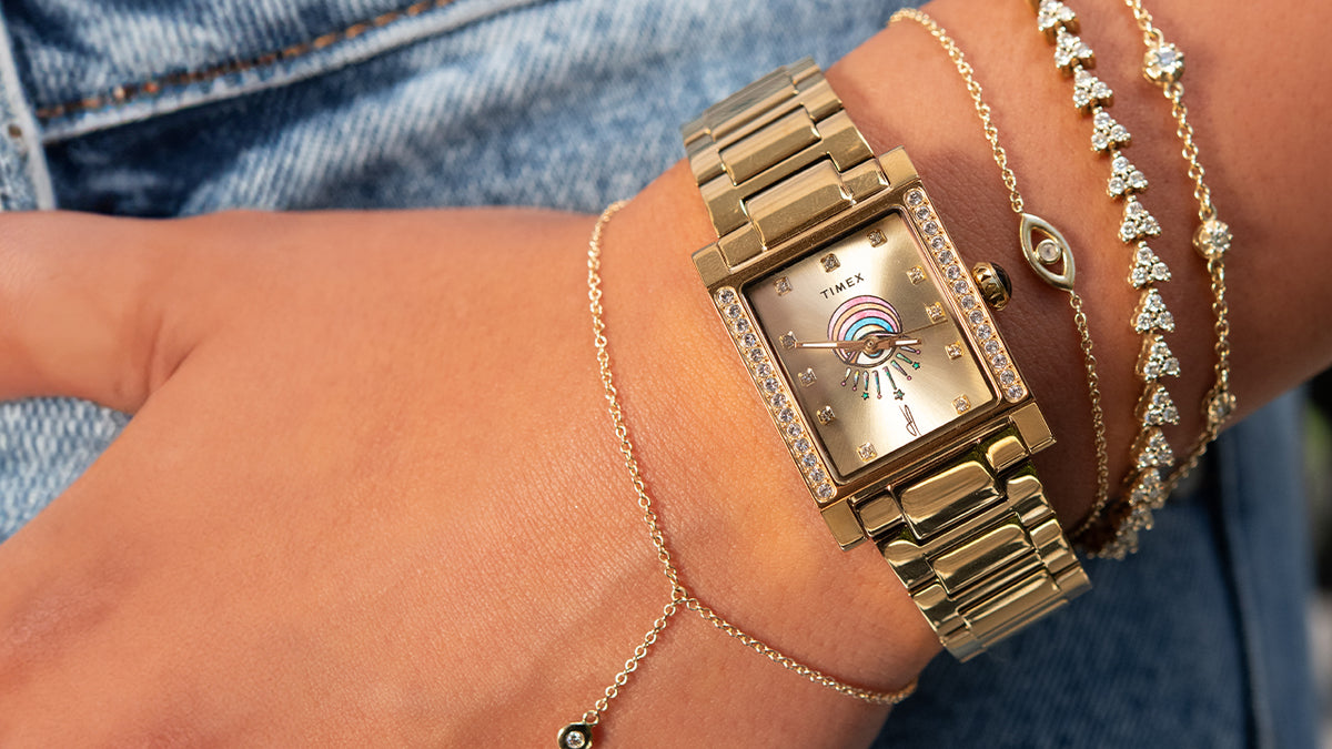 Gold-tone Timex x Jacquie Aiche Éclat watch worn with layered gold and crystal bracelets.