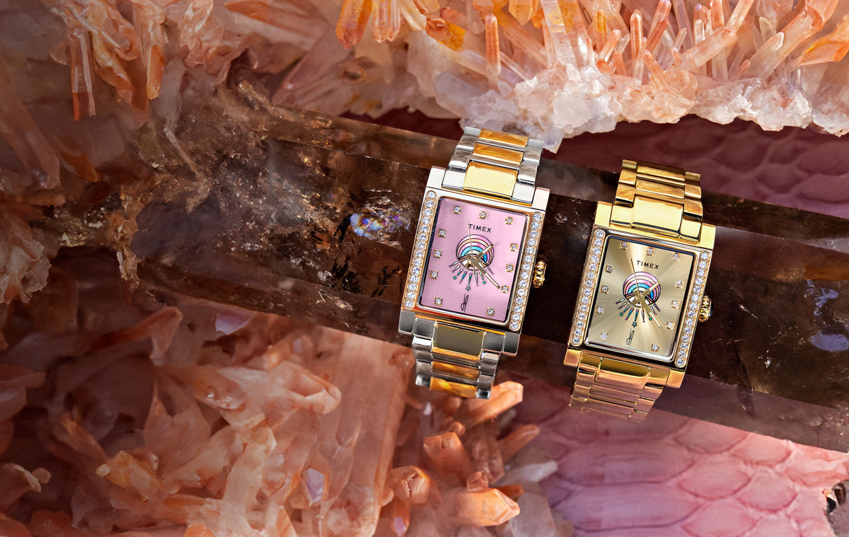Two Timex x Jacquie Aiche Éclat watches with pink and champagne eye-motif dials displayed among pink crystals.