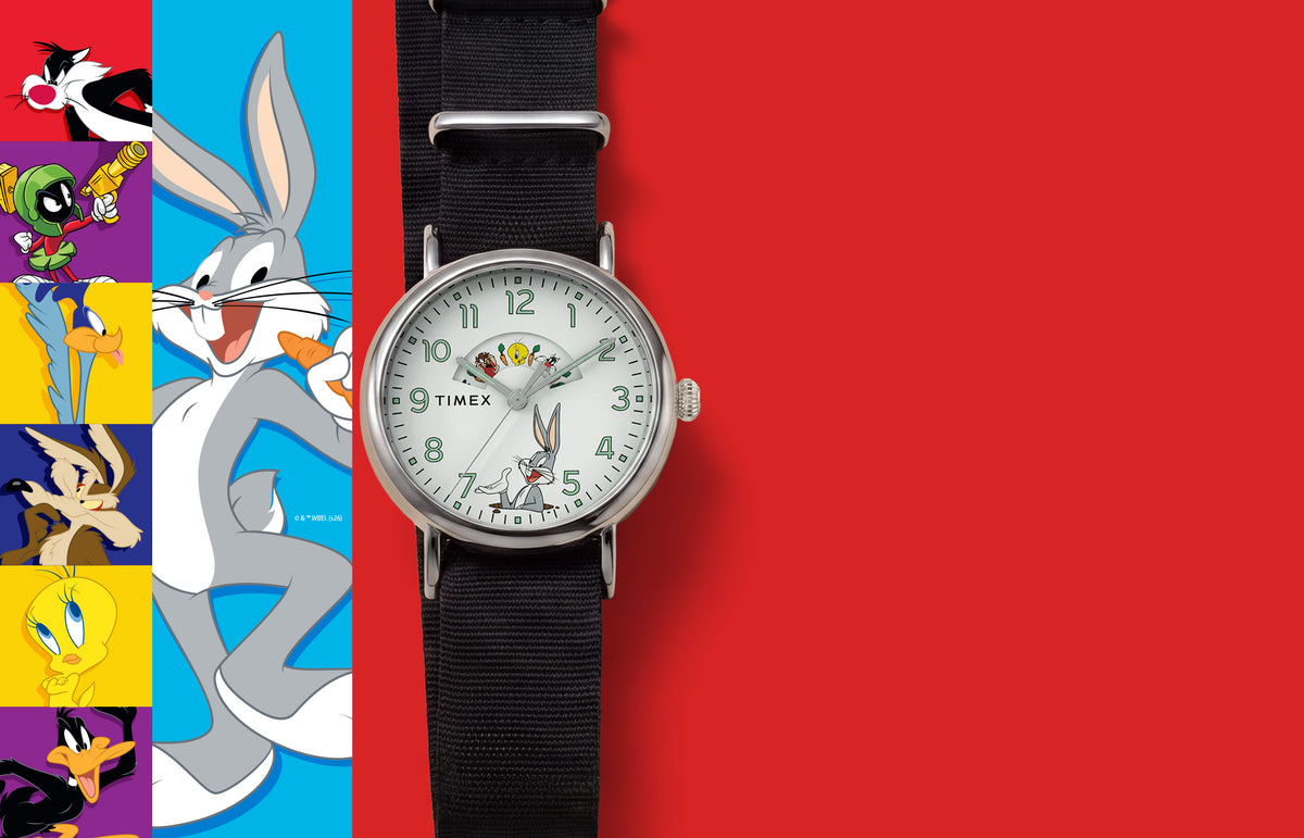 TW2Y99100 Looney Tunes Bugs and Friends x Timex Standard watch with a white character dial and black fabric strap beside colorful Looney Tunes artwork.