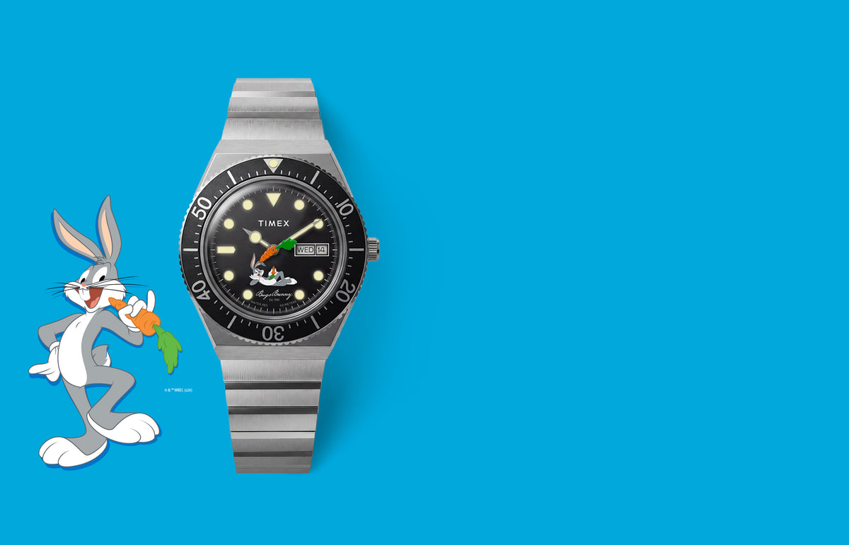 TW2Y99200 Looney Tunes Bugs Bunny x Timex M79 Automatic Signature Edition watch with a black dial and silver-tone bracelet beside Bugs Bunny artwork on blue.