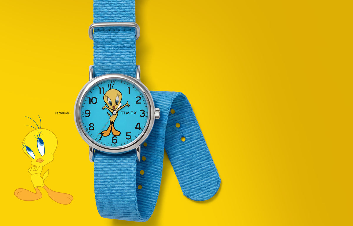 TW2Y99500 Looney Tunes Tweety Bird x Timex Weekender watch with a blue character dial and blue fabric strap beside Tweety artwork on yellow.