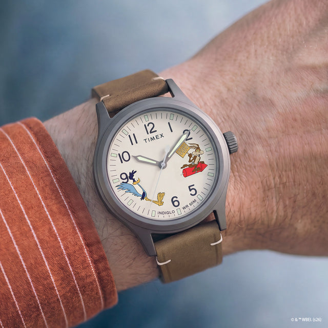 Reloj TW2Y99400 Road Runner & Wile E. Coyote x Timex Expedition® Scout, llevado en la muñeca, con esfera color crema, caja gris y correa de piel marrón.