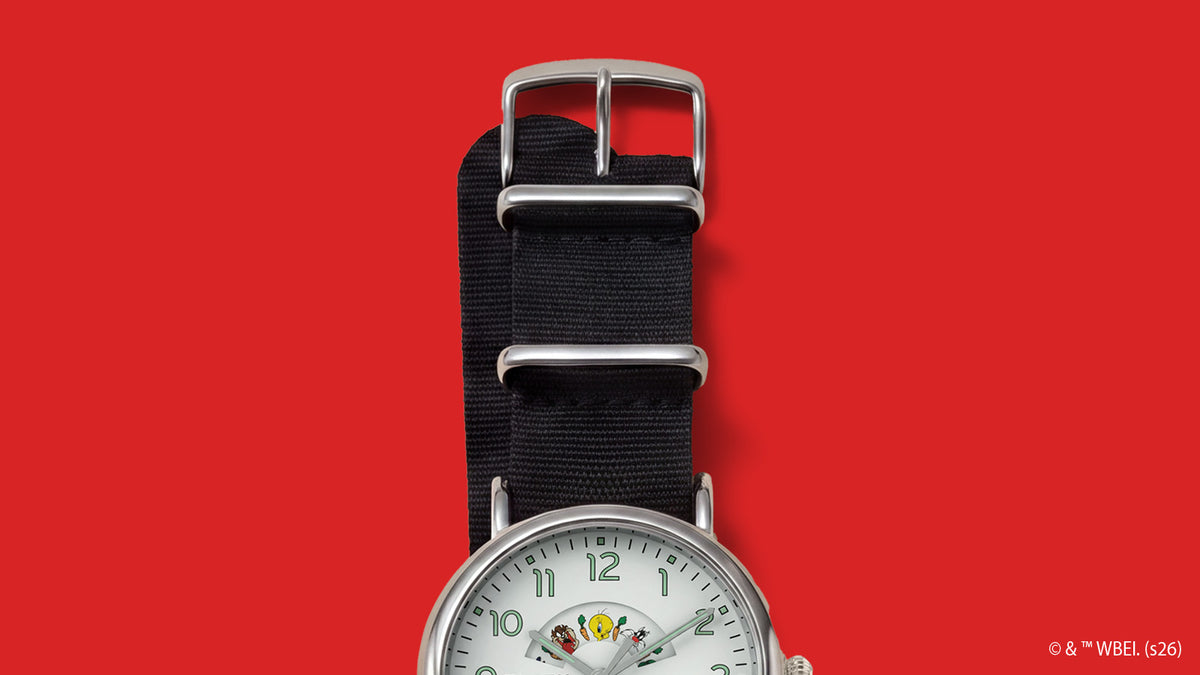 TW2Y99100 Black woven strap with a silver-tone buckle and keepers on the Looney Tunes Bugs and Friends x Timex Standard watch against red.