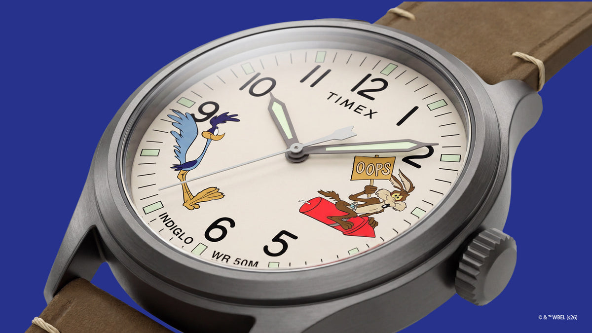 TW2Y99400 Vista en ángulo del reloj Road Runner & Wile E. Coyote, en la que se aprecia su esfera de color crema con motivos de los personajes, los números negros, la caja gris y la correa de piel marrón.