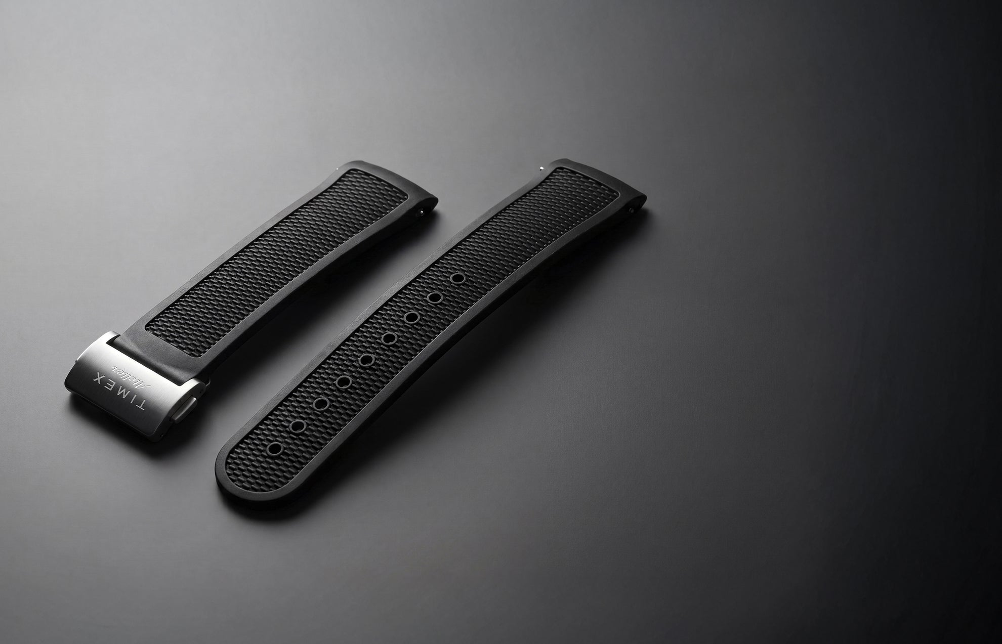 Timex Atelier NBR synthetic rubber strap in black with a textured finish and stainless-steel clasp, displayed against a dark background.