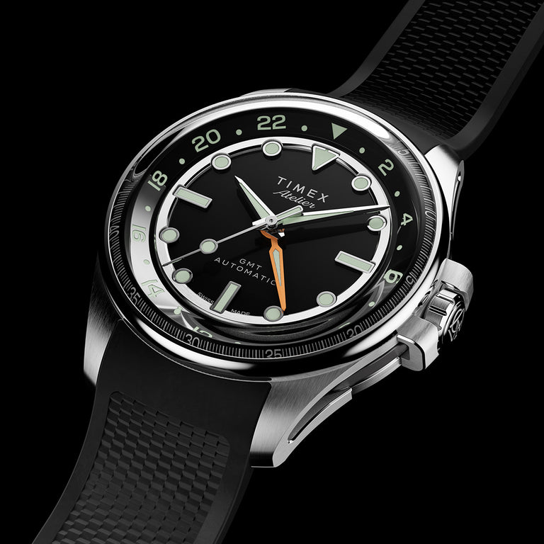Reloj automático Timex Atelier GMT con esfera negra, índices luminosos, manecilla GMT naranja y correa de caucho negro con textura.
