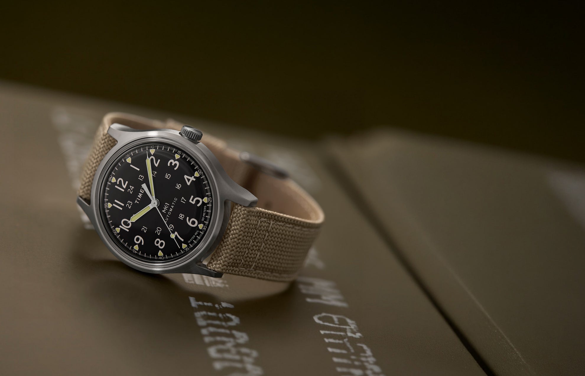 MK1® Automatic watch with a black dial, brushed silver-tone case, and tan fabric strap resting on military-style equipment.