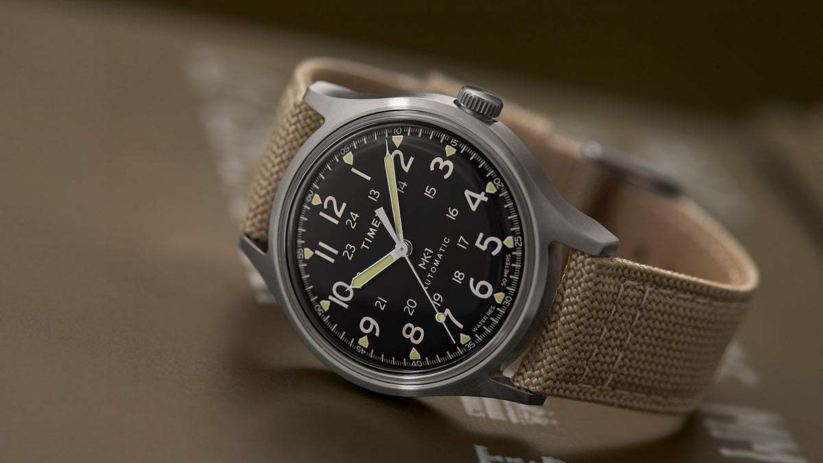 Close-up of the MK1® Automatic watch with a black dial, stainless-steel case, and tan fabric strap.