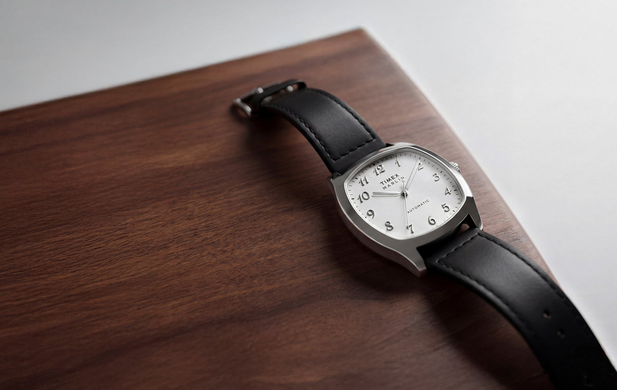 TW2Y84300 Marlin® Chatham Automatic watch with a silver-tone cushion-shaped case, white dial, and black leather strap resting on a wooden surface.