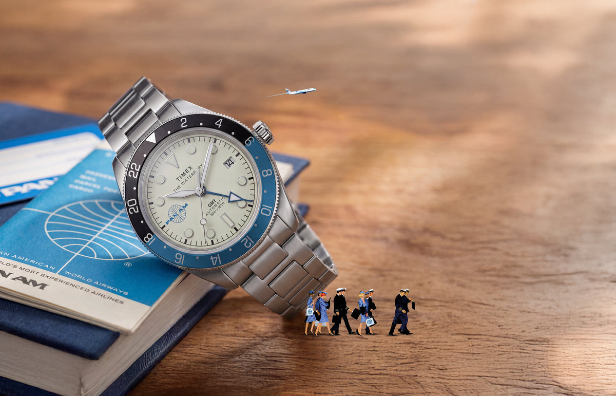TW2Y94300 Pan Am x Timex Waterbury Automatic GMT watch beside a Pan Am travel book with miniature airline crew and airplane details.