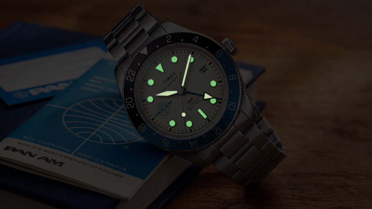 TW2Y94300 Pan Am x Timex Waterbury Automatic GMT watch glowing beside a vintage Pan Am travel book.