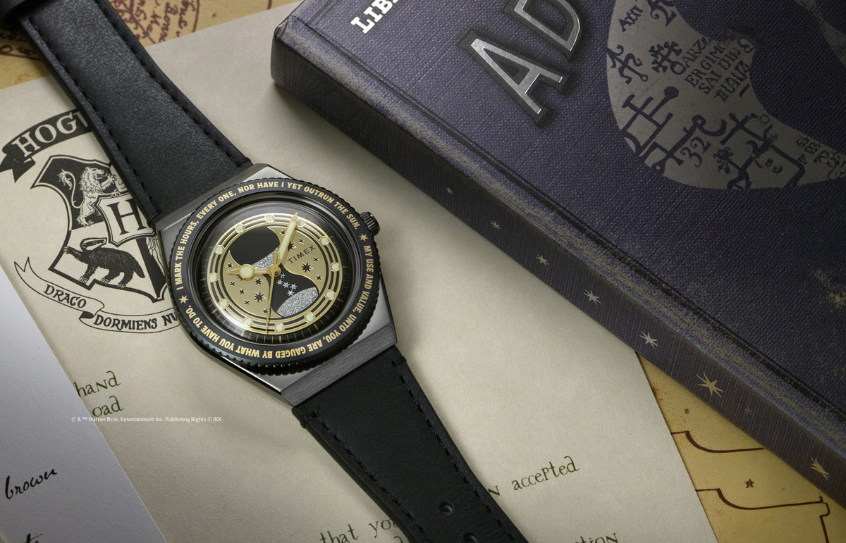 TW2Y76000 Black watch with a gold hourglass and stars on the dial, laid across Hogwarts-themed papers beside a book.