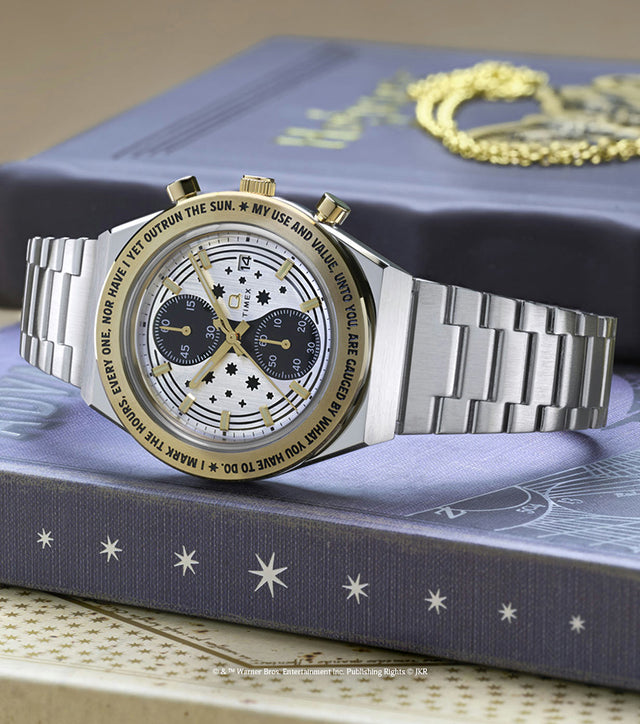 TW2Y75900 Silver-tone chronograph watch with a star-patterned dial and gold-tone bezel displayed beside a gold Time-Turner pendant on a book.