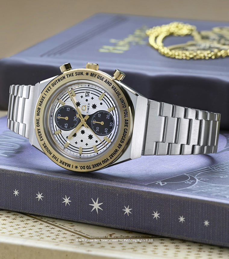 TW2Y75900 Silver-tone chronograph watch with a star-patterned dial and gold-tone bezel displayed beside a gold Time-Turner pendant on a book.