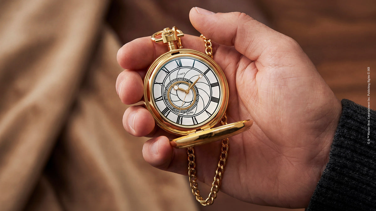 TW2Y99600 Hand holding the open gold-tone pocket watch, showing its white dial and hanging chain.