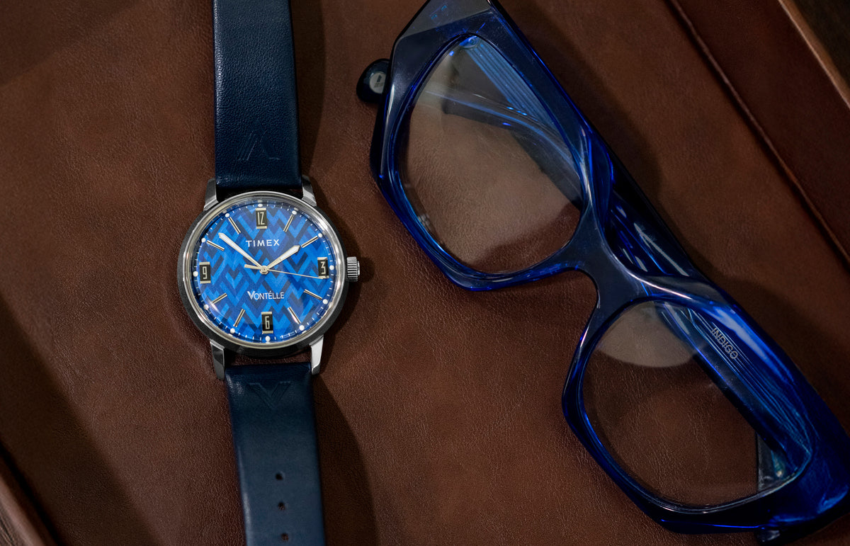TW2Y73800 Blue patterned dial watch with a navy strap laid beside blue eyeglasses on a brown surface.
