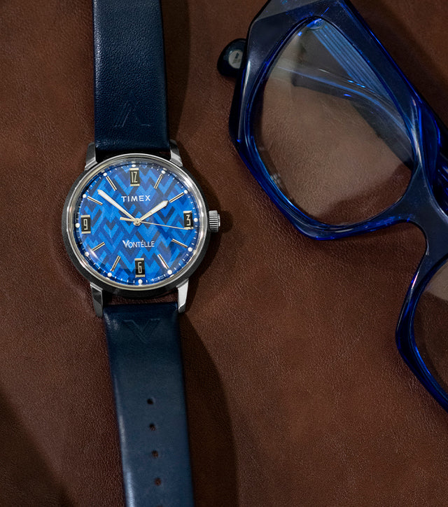 TW2Y73800 Blue patterned dial watch with a navy strap laid beside blue eyeglasses on a brown surface.