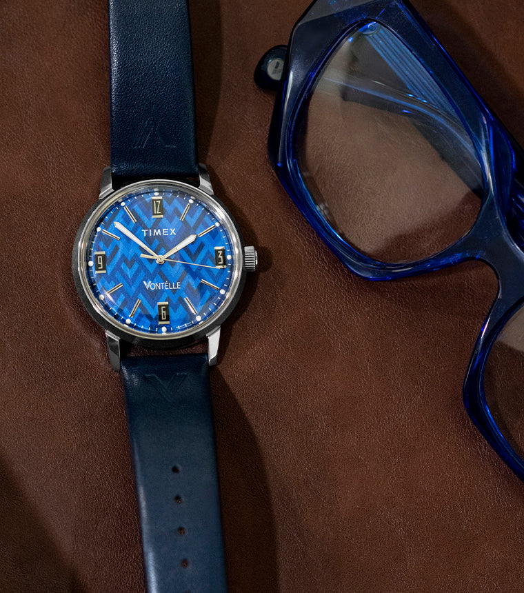 TW2Y73800 Blue patterned dial watch with a navy strap laid beside blue eyeglasses on a brown surface.
