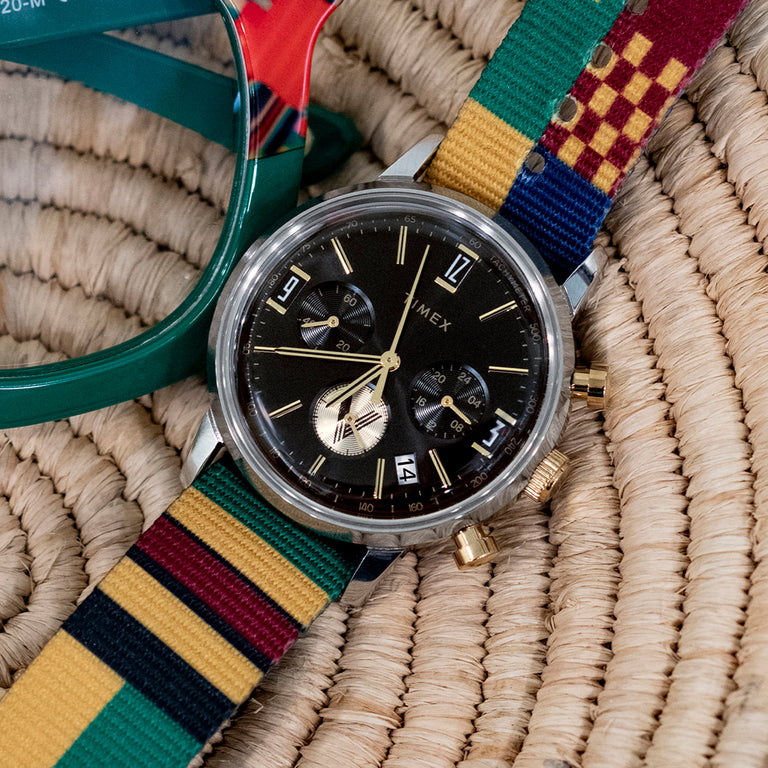 Reloj TWG069100 con esfera negra y correa a rayas de colores, expuesto sobre una superficie tejida junto a unas gafas verdes.
