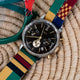Reloj TWG069100 con esfera negra y correa a rayas de colores, expuesto sobre una superficie tejida junto a unas gafas verdes.