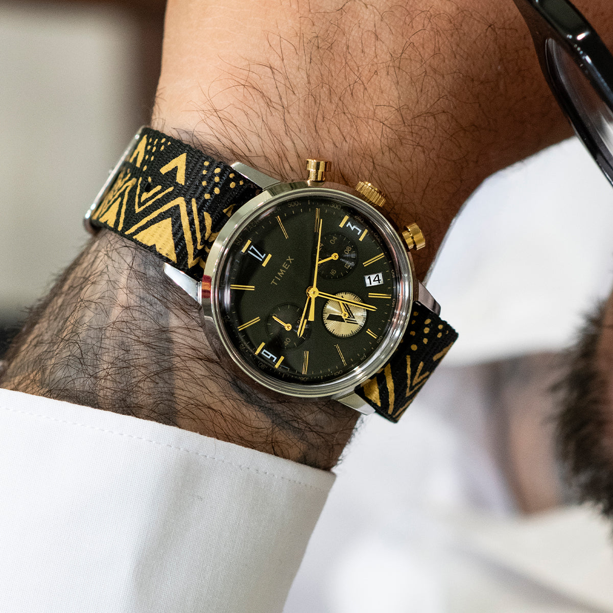 TWG069100 Primer plano de un reloj de esfera negra con una correa con motivos en negro y dorado, puesto en la muñeca.