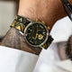 TWG069100 Primer plano de un reloj de esfera negra con una correa con motivos en negro y dorado, puesto en la muñeca.