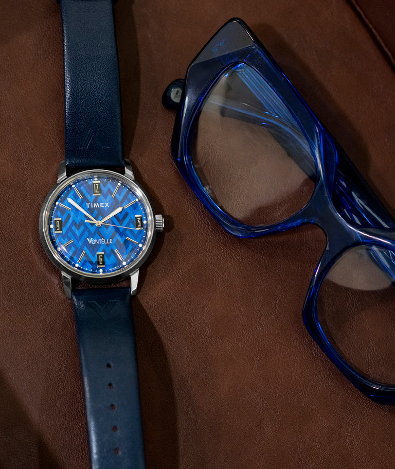 TW2Y73800 Vontélle x Timex Marlin watch with a blue geometric dial, silver-tone case and navy strap beside blue eyeglasses on a brown leather tray.