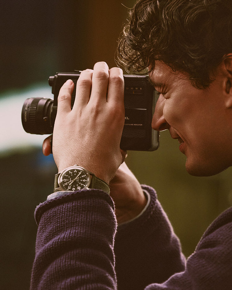 Gifts for Him - Man using a vintage movie camera while wearing a Timex watch with a black dial and green strap.