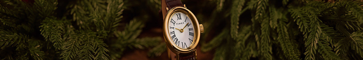 Favoritos navideños: reloj Timex con caja ovalada en tono dorado, esfera blanca con números romanos y correa de piel marrón, rodeado de ramas de hoja perenne.