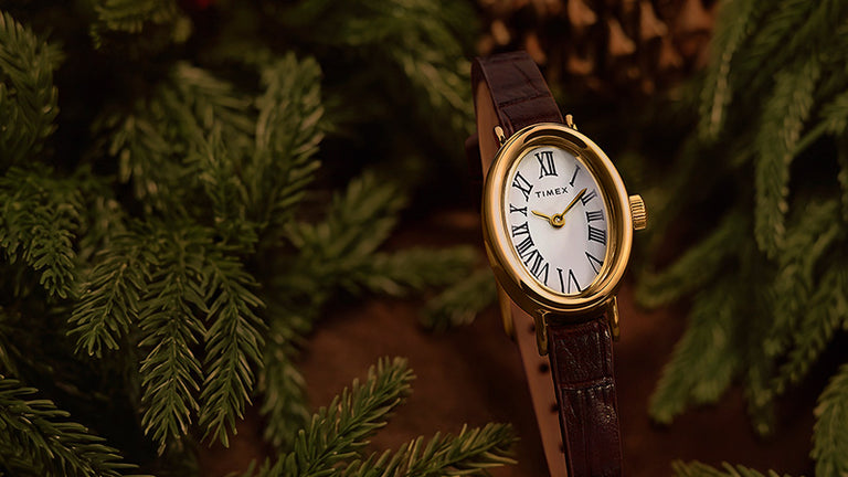 Favoritos navideños: reloj Timex con caja ovalada en tono dorado, esfera blanca con números romanos y correa de piel marrón, rodeado de ramas de hoja perenne.