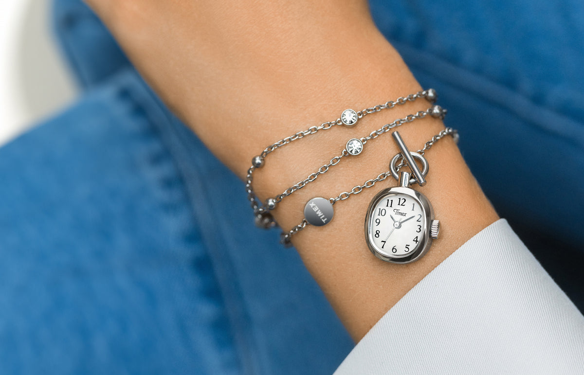 TW6A17600 Primer plano del collar-reloj Cavatina® Mini colocado en la muñeca, junto a un puño blanco, sobre un fondo de vaquero azul.