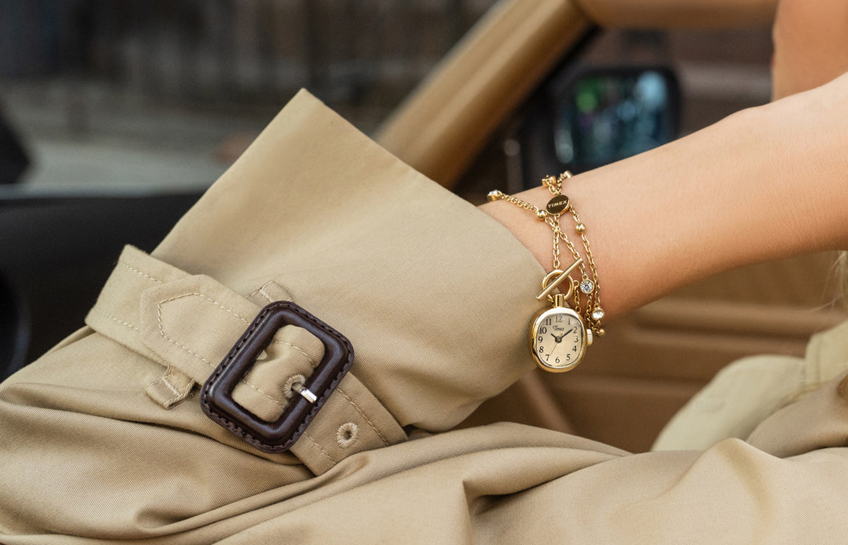 TW6A17700 Primer plano de un collar con reloj Cavatina® Mini en tono dorado que rodea una muñeca, junto al puño de una gabardina beige.