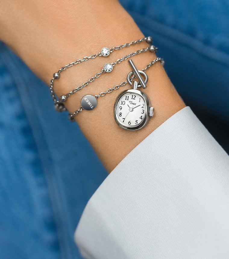 TW6A17600 Primer plano del collar-reloj Cavatina® Mini colocado en la muñeca, junto a un puño blanco, sobre un fondo de vaquero azul.