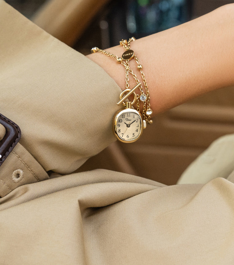 TW6A17700 Primer plano de un collar con reloj Cavatina® Mini en tono dorado que rodea una muñeca, junto al puño de una gabardina beige.