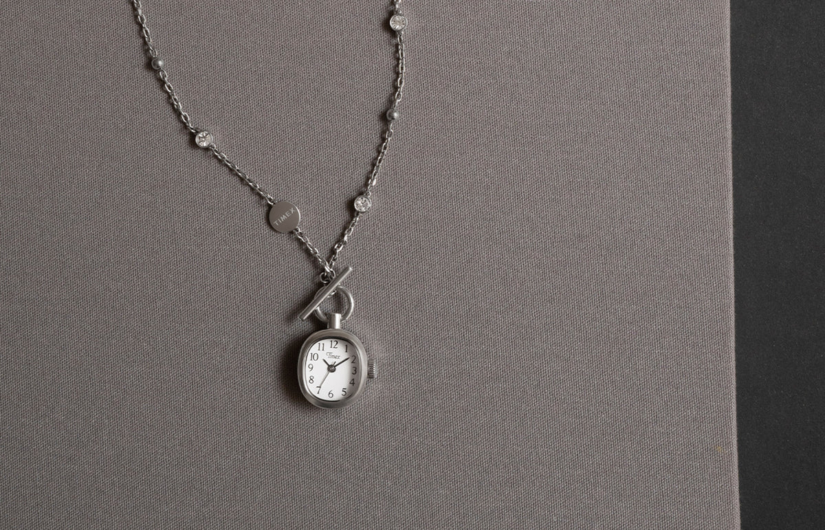 TW6A17600 Collar Cavatina® Mini con un colgante en forma de reloj de esfera ovalada blanca y una cadena plateada con piedras transparentes, presentado sobre tela gris.