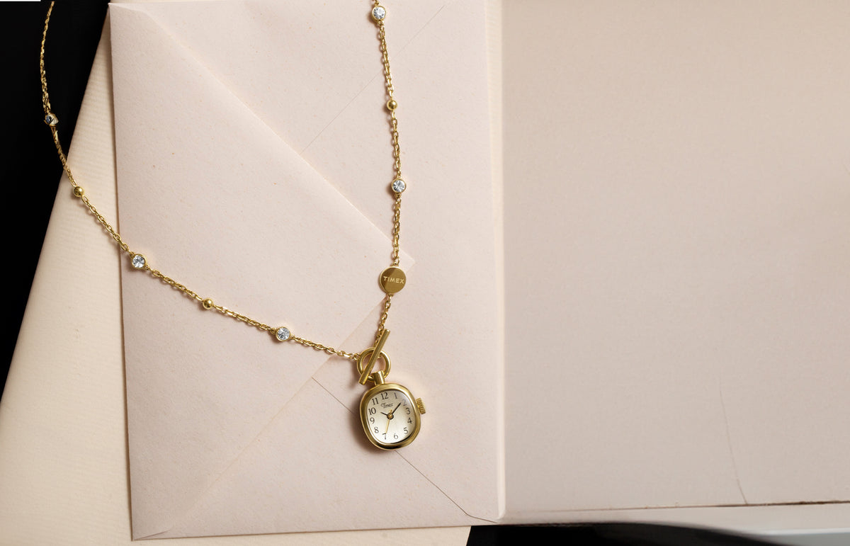 TW6A17700: collar con reloj Cavatina® Mini en tono dorado, con piedras transparentes y cierre de palanca, presentado sobre papel de carta color crema.