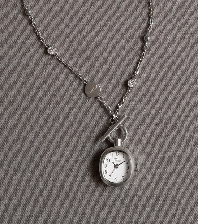 TW6A17600 Collar Cavatina® Mini con un colgante en forma de reloj de esfera ovalada blanca y una cadena plateada con piedras transparentes, presentado sobre tela gris.