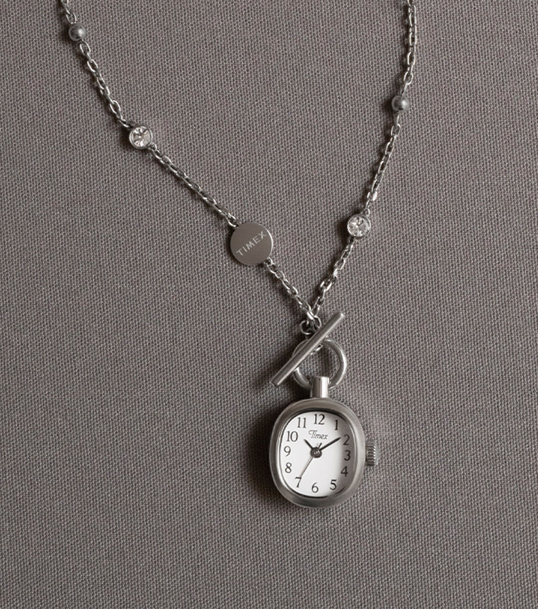 TW6A17600 Collar Cavatina® Mini con un colgante en forma de reloj de esfera ovalada blanca y una cadena plateada con piedras transparentes, presentado sobre tela gris.