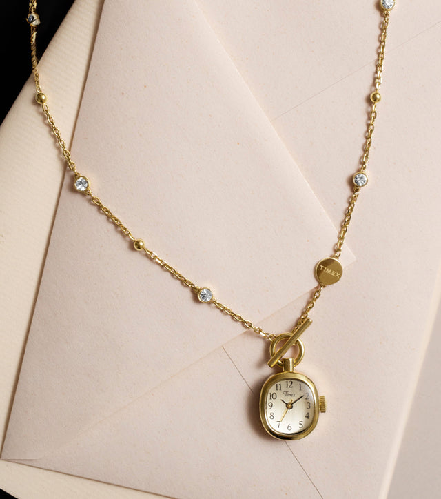 TW6A17700: collar con reloj Cavatina® Mini en tono dorado, con piedras transparentes y cierre de palanca, presentado sobre papel de carta color crema.