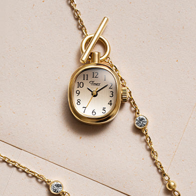 TW6A17700 Colgante para reloj Cavatina® Mini en tono dorado con números arábigos negros, junto a una cadena con piedras transparentes sobre papel color crema.