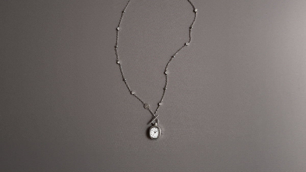 TW6A17600 Collar Cavatina® Mini de acero inoxidable con un colgante en forma de reloj, cierre de palanca y cadena con piedras transparentes, presentado sobre una tela gris.