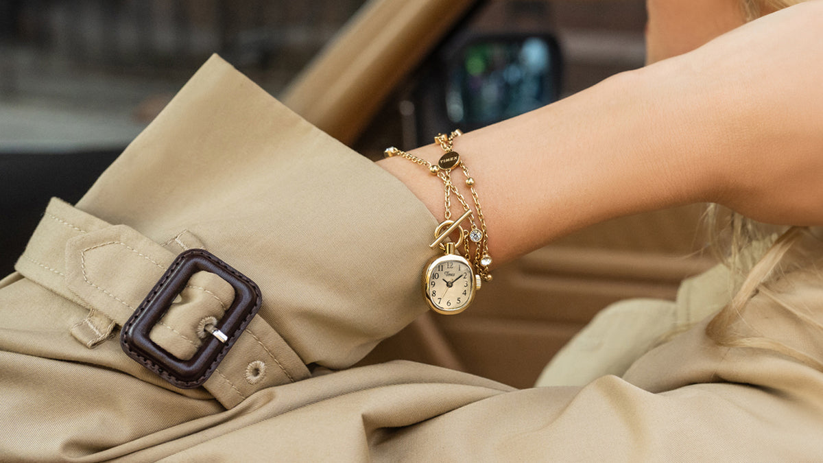TW6A17700: collar Cavatina® Mini en tono dorado enrollado alrededor de una muñeca, con el colgante en forma de reloj junto al puño de una gabardina beige.