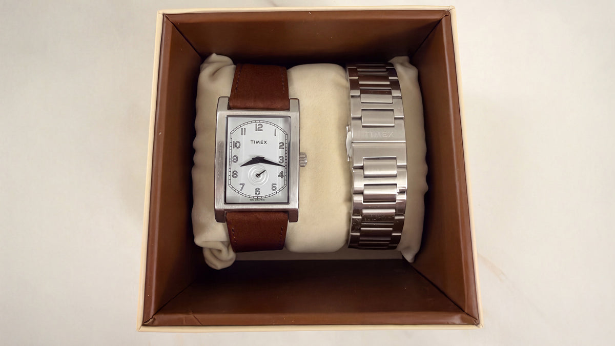 Reloj rectangular TWG069000 WM Brown x Timex con brazalete plateado y una correa adicional de piel marrón, presentado en una caja abierta.