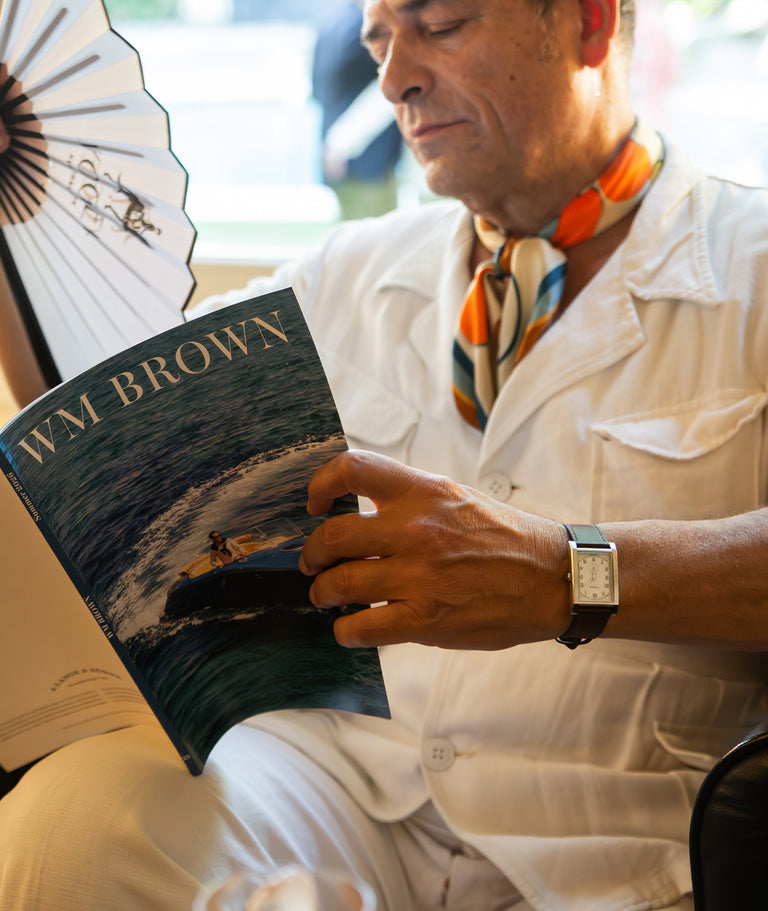 TWG069000: Hombre leyendo una revista de WM Brown y sosteniendo un abanico plegable, que lleva un reloj rectangular de WM Brown x Timex con esfera color crema y correa de cuero oscuro.