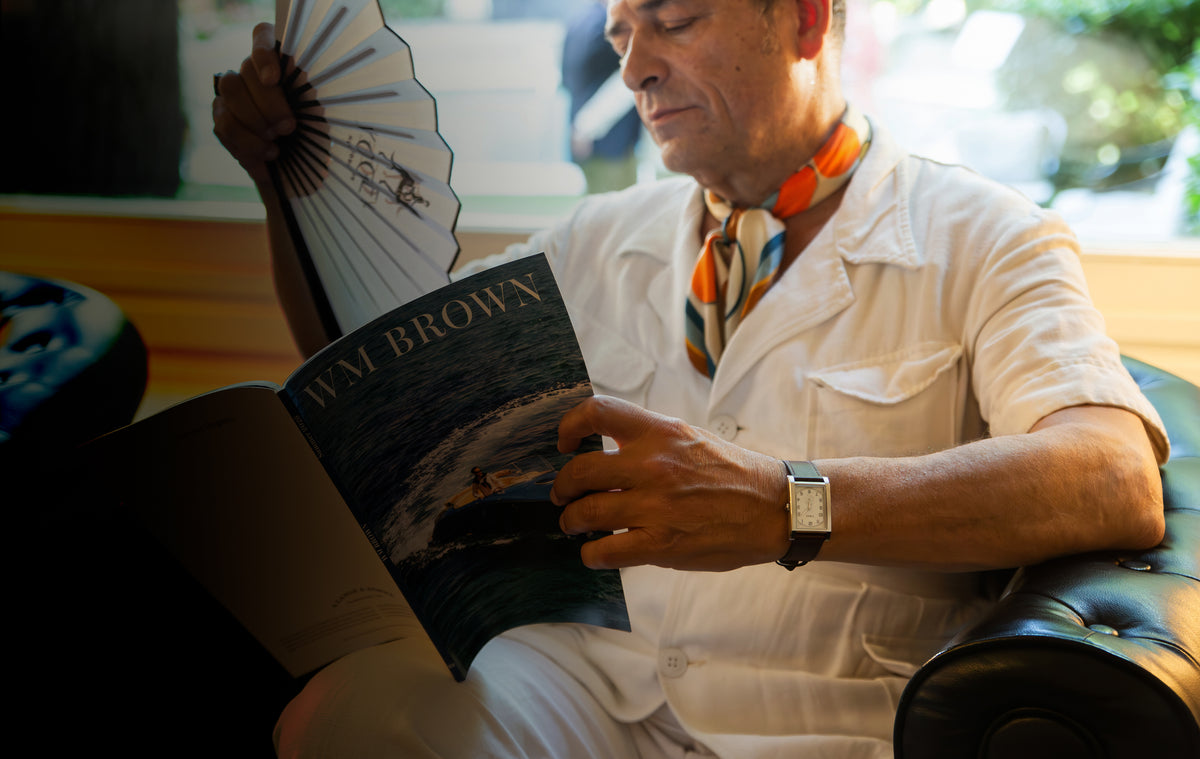 TWG069000 Man reading WM Brown magazine and holding a folding fan while wearing a rectangular WM Brown x Timex watch with a cream dial and dark leather strap.