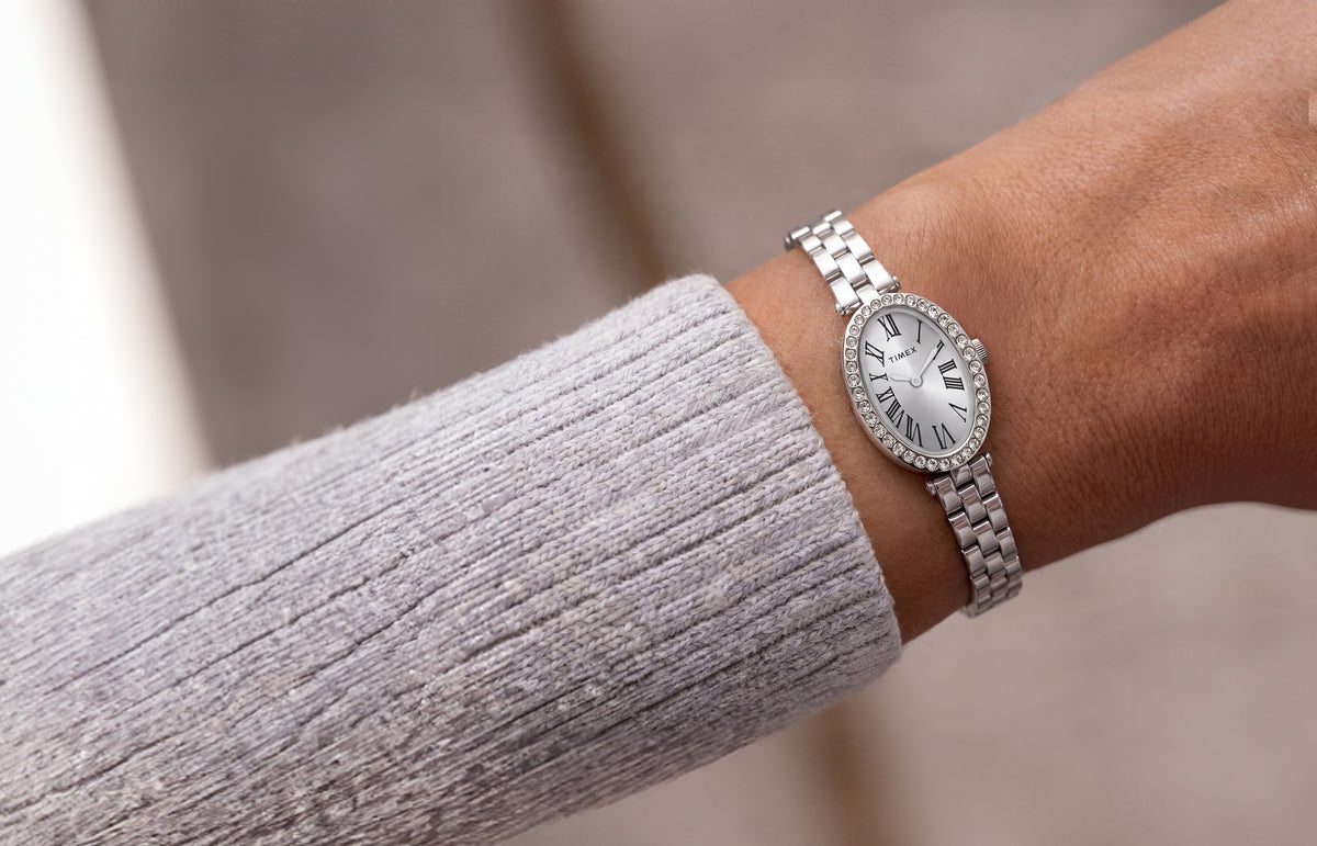 Reloj Timex Cavatina® Crystals TW2Y90000 con brazalete de eslabones en tono plateado y esfera ovalada con números romanos, lucido junto a una manga de punto gris claro.