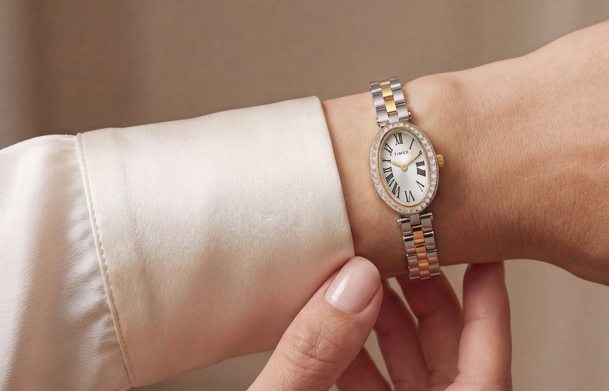 TW2Y90200: Reloj Timex Cavatina® Crystals en dos tonos, con bisel ovalado adornado con cristales y esfera con números romanos, lucido junto al puño de una blusa de color marfil.