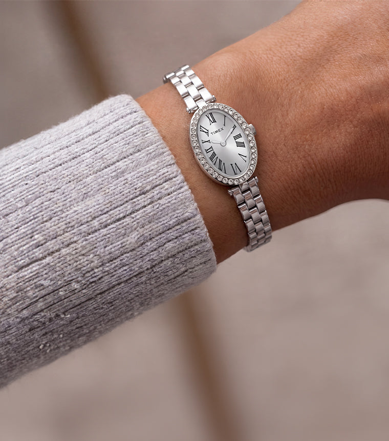 Reloj Timex Cavatina® Crystals TW2Y90000 con brazalete de eslabones en tono plateado y esfera ovalada con números romanos, lucido junto a una manga de punto gris claro.